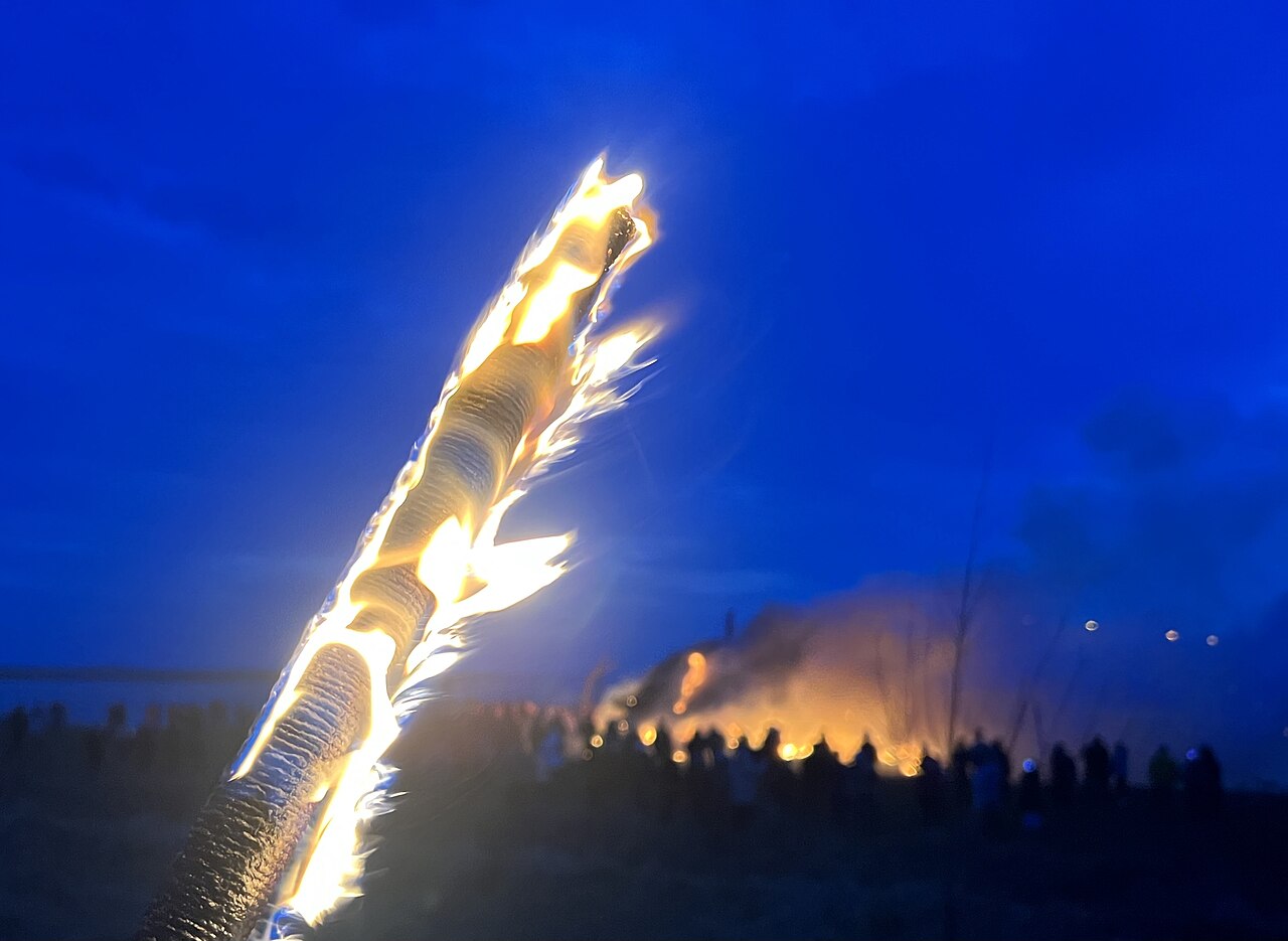 Ein hoch aufloderndes Biike-Feuer erhellt die Dunkelheit, davor stehen Zuschauer als Silhouetten.
